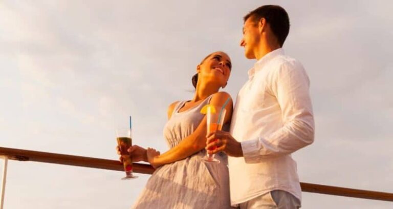 A couple enjoying drinks while leaning against a railing during sunset, smiling at each other.