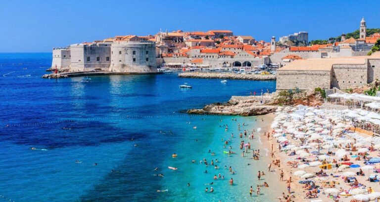 A sunny coastline scene with people on a beach, clear blue water, and the historic city of Dubrovnik featuring orange rooftops in the background.