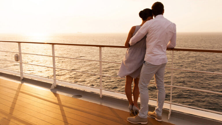 A couple stands on a ship's deck, watching the sunset over the ocean.