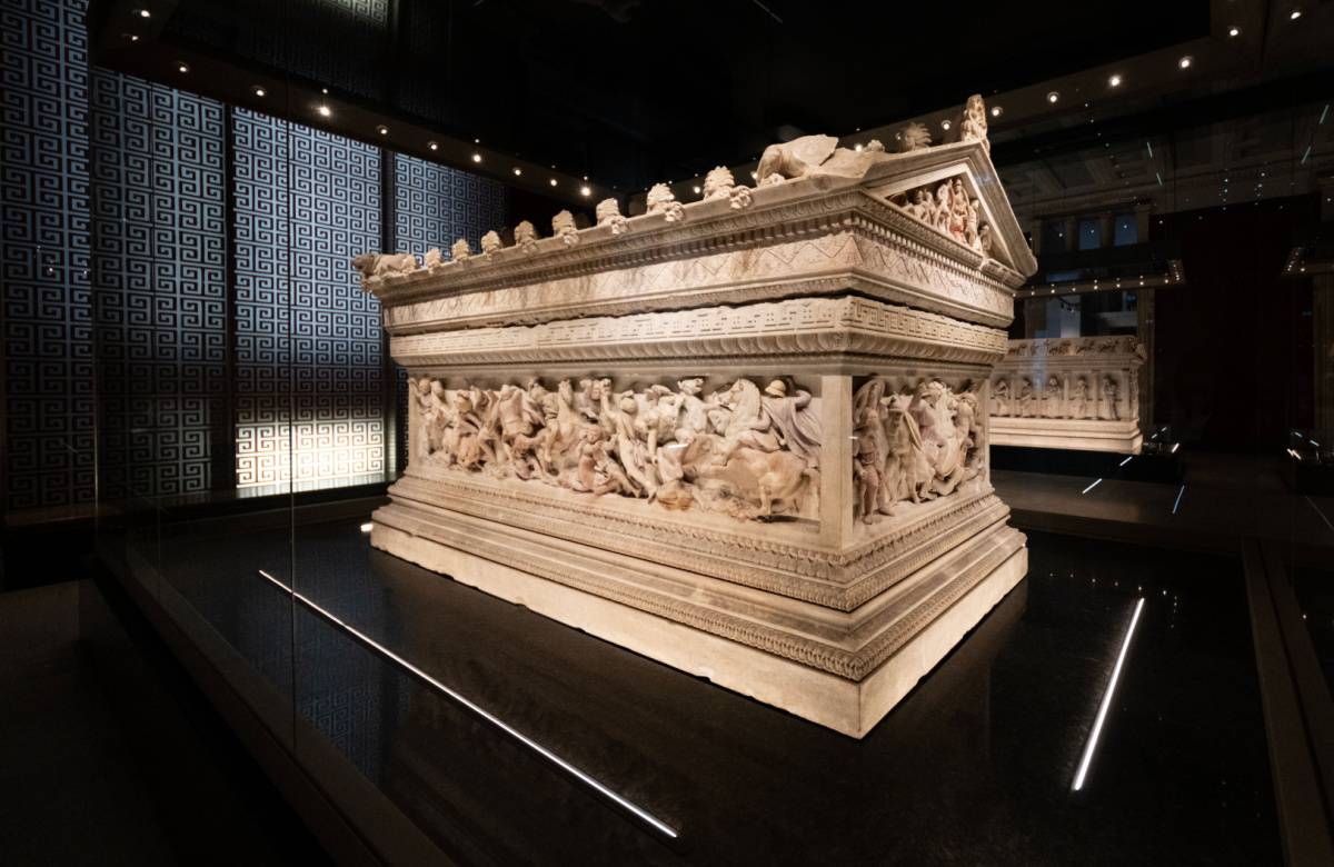 The intricate Alexander Sarcophagus displayed inside the Istanbul Archaeology Museum, beautifully lit and showcased against a patterned backdrop, highlighting the detailed carvings of figures in high relief.