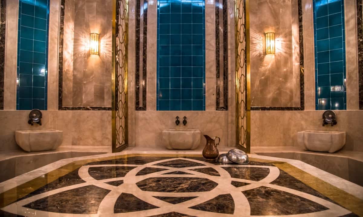 An ornate Turkish bath interior with marble floors and walls, featuring two sinks and a decorative tiled wall, with traditional bath items on display.