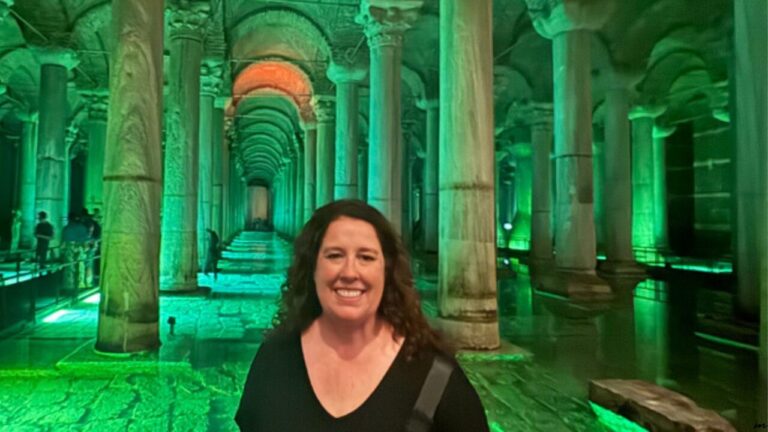 Smiling person standing inside a large, ornately columned underground space with green lighting.