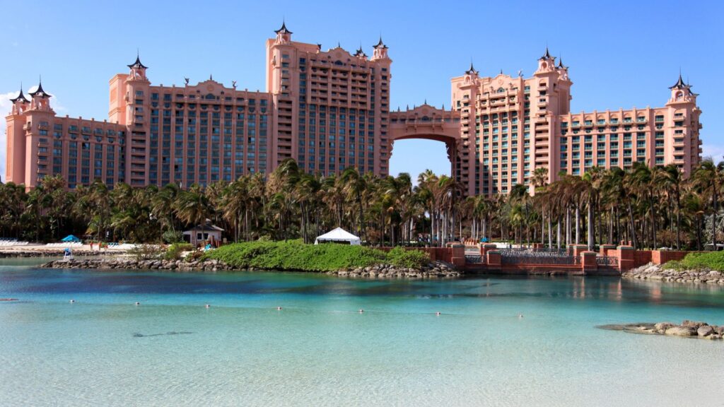 Pink luxurious resort with towers by a turquoise lagoon, surrounded by palm trees under a clear blue sky.