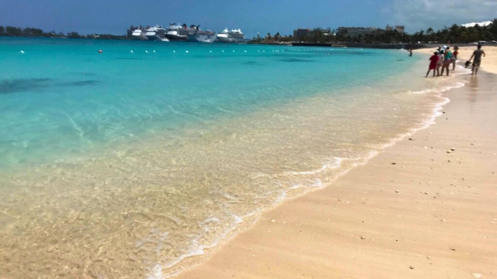 A serene beach with turquoise water, people walking along the shore, and cruise ships visible in the distance.