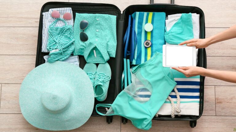 Open suitcase packed with turquoise clothes, striped towel, hat, and sunglasses. A person is holding a blank tablet above it.