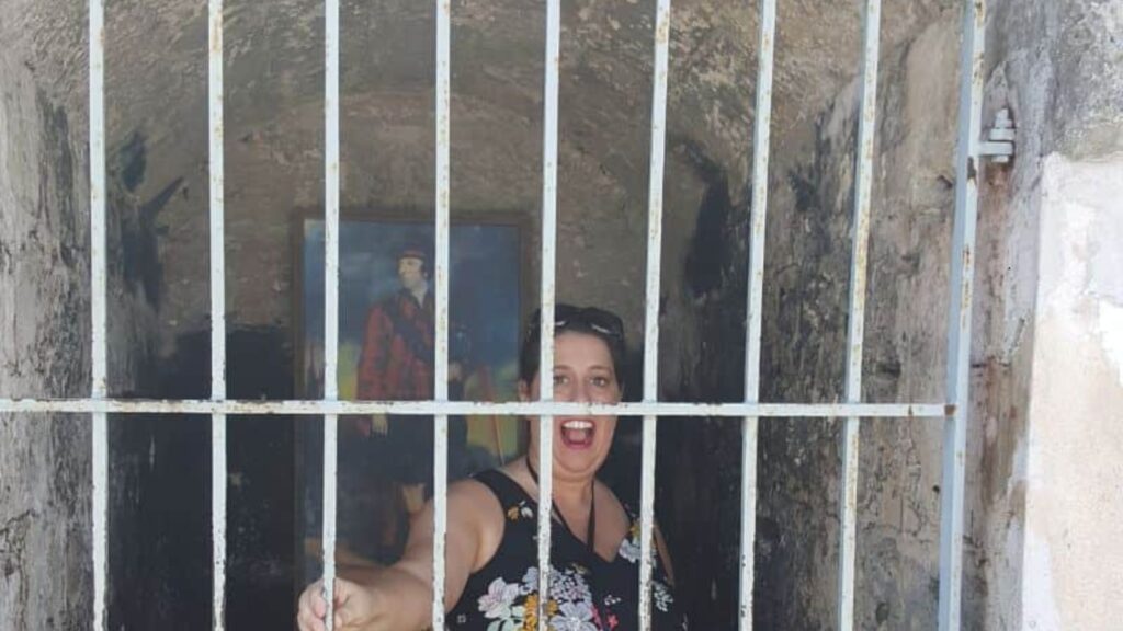 A woman smiling and posing behind bars in a dimly lit stone cell, with a painting visible in the background.