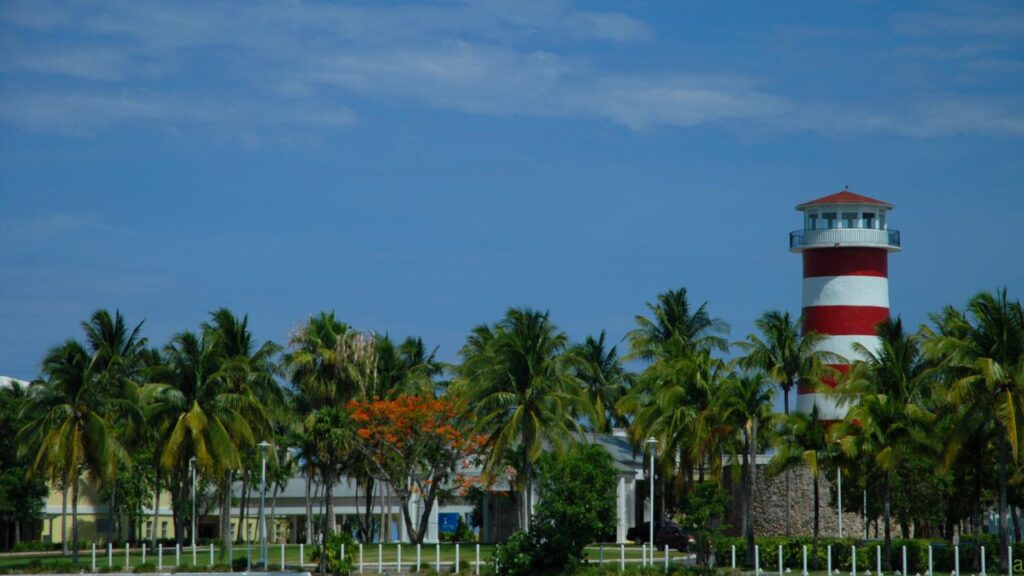 A red and white striped lighthouse stands among palm trees against a blue sky.