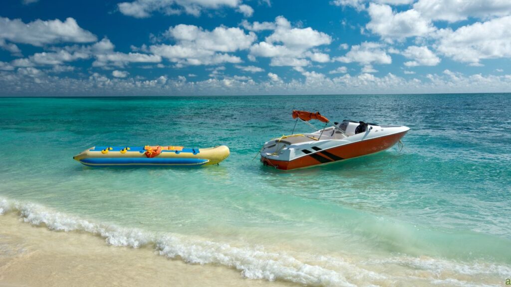 A speedboat and an inflatable banana boat float on calm, clear turquoise water near a sandy beach under a partly cloudy sky.