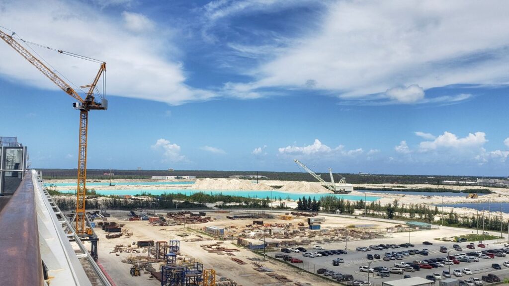 Wide view of construction site with cranes, sand piles, parking lot, and distant water under a partly cloudy sky.