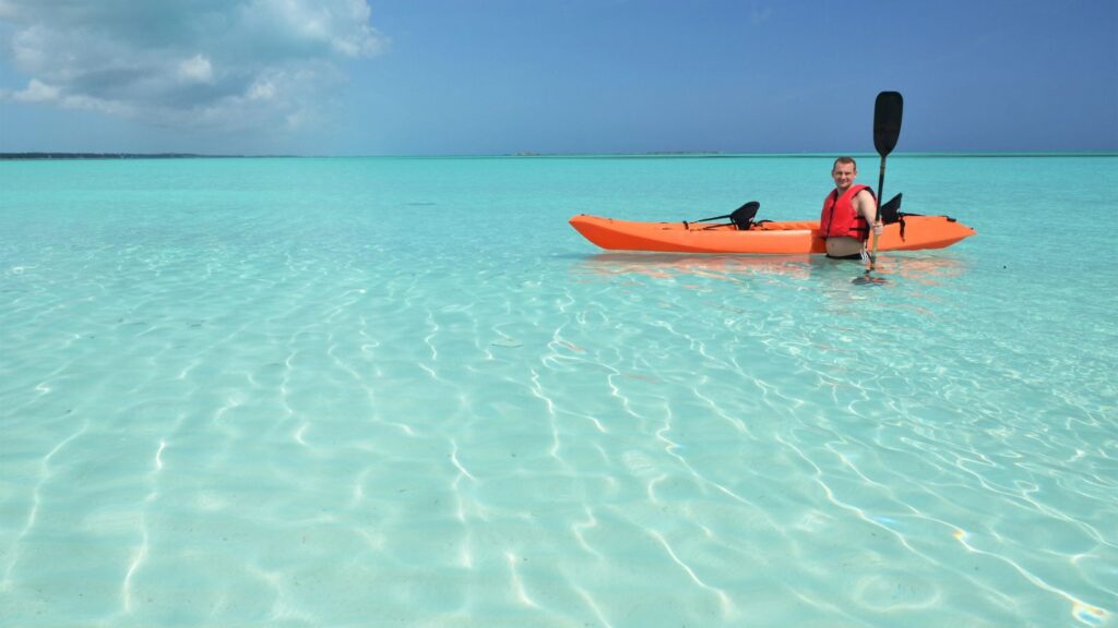 Man in a red life jacket sits in an orange kayak on clear turquoise waters under a sunny sky.