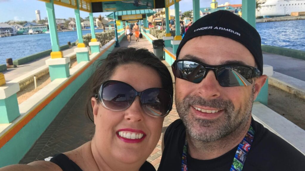 A man and woman wearing sunglasses smile together on a sunny waterfront path with colorful buildings in the background.