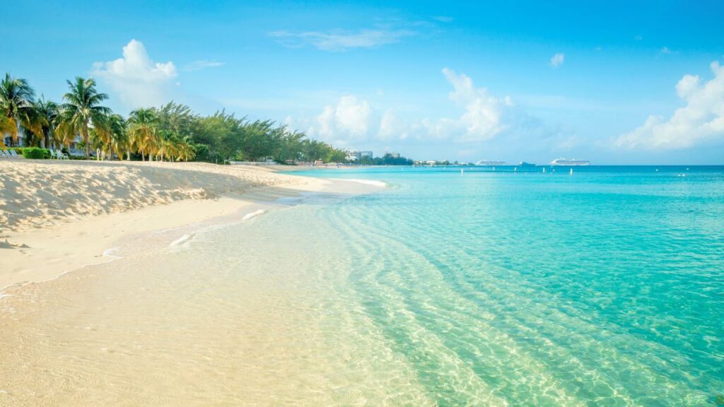 A serene beach with crystal-clear turquoise water, white sandy shore, palm trees, and calm clouds in the blue sky.