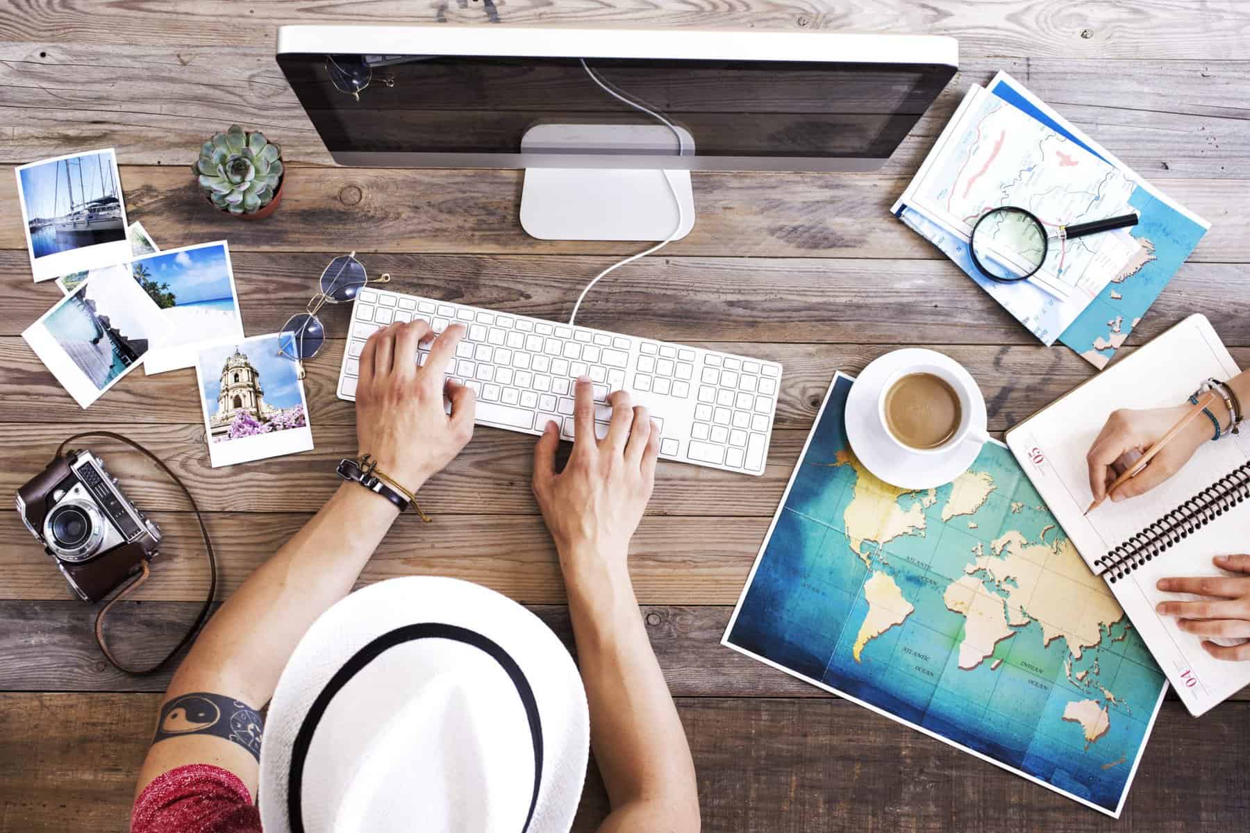 Top view of a person typing on a keyboard with travel photos, maps, a camera, and coffee on a wooden table.