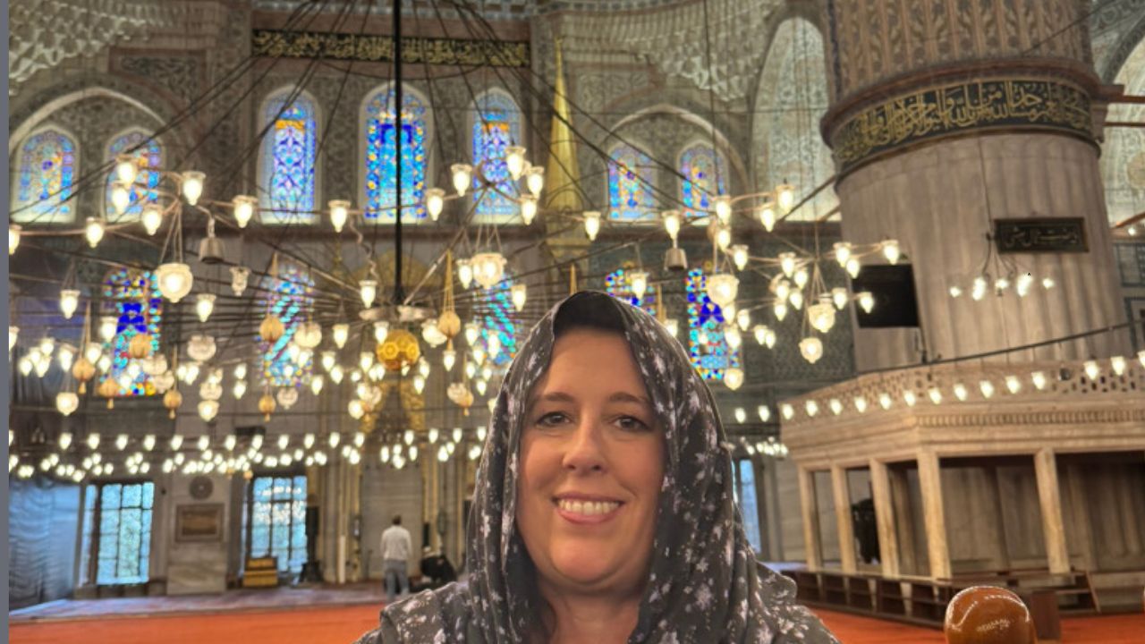 A woman in a headscarf smiles inside a mosque adorned with stained glass windows and numerous hanging lights.