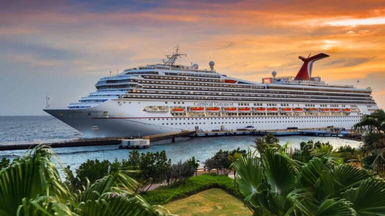 A large cruise ship docked at port with a sunset sky in the backdrop, surrounded by tropical greenery.