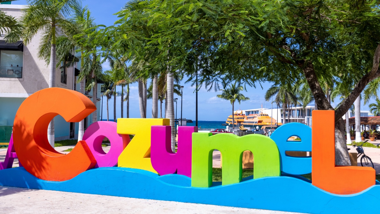 Multicolored "Cozumel" sign with palm trees and buildings in the background on a sunny day.