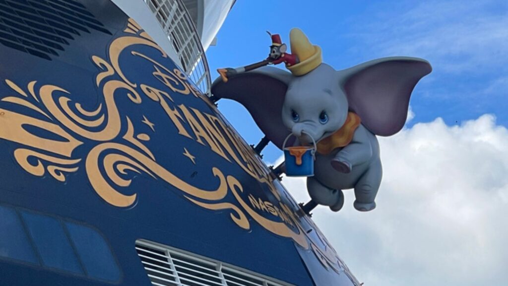 A figure of Dumbo the Flying Elephant is attached to the exterior of a Disney-themed cruise ship against a blue sky.