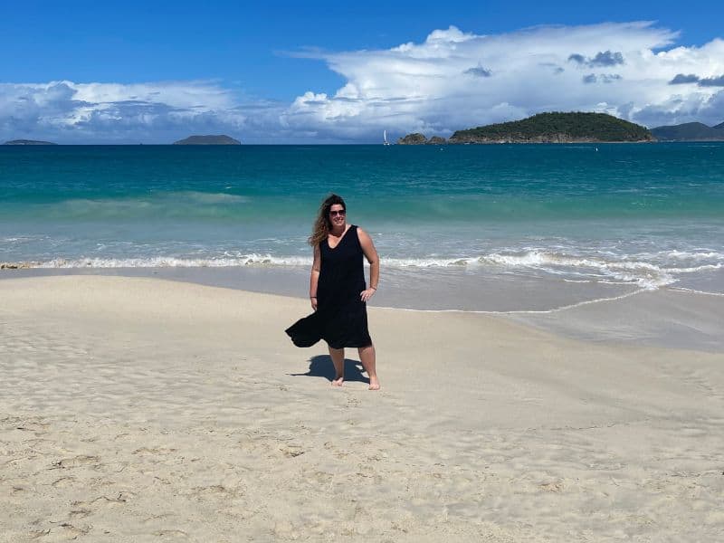 Person in black dress stands on sandy beach with turquoise water, islands in background, and partially cloudy sky.