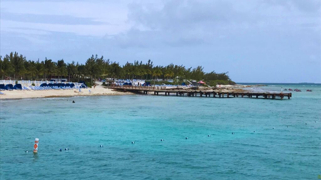 A wooden pier extends into the turquoise sea with a sandy beach and sun loungers under a cloudy sky in the background.