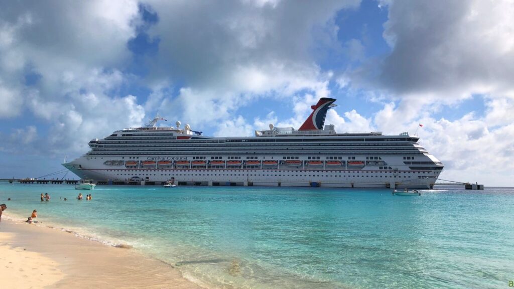 A cruise ship docked near a beach with clear blue water and people swimming in the foreground under a partly cloudy sky.