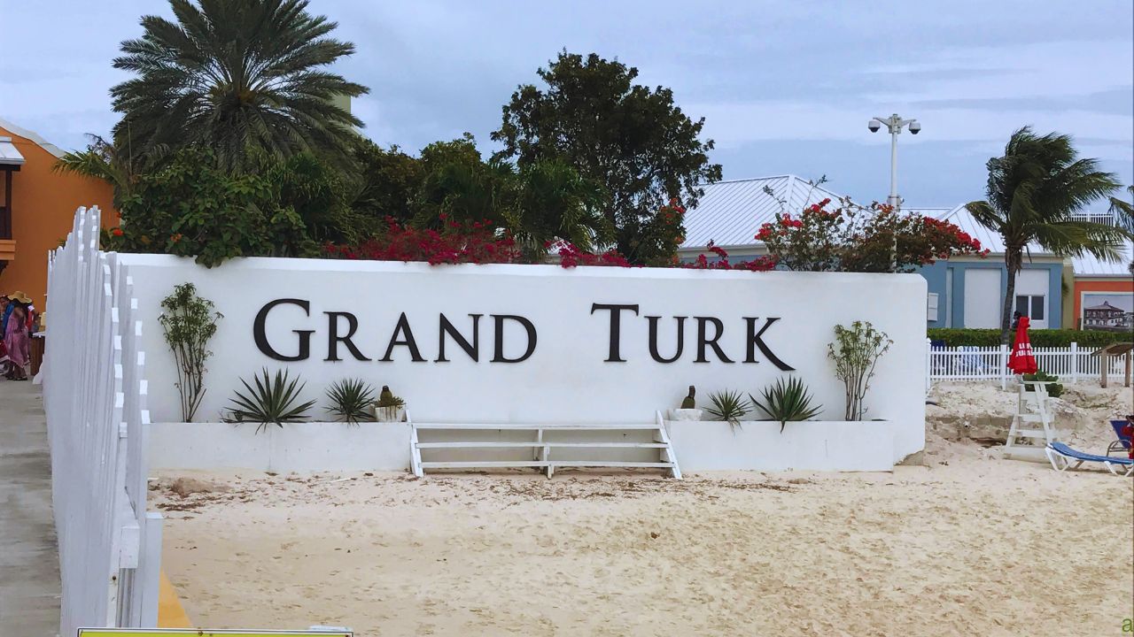 A white sign on a sandy beach reads "Grand Turk" with tropical plants and palm trees in the background.