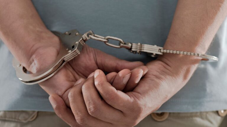 Close-up of a person's hands behind their back in handcuffs, wearing a light blue shirt.