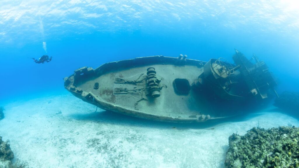 A scuba diver explores an underwater shipwreck on the ocean floor, with clear blue water in the background.