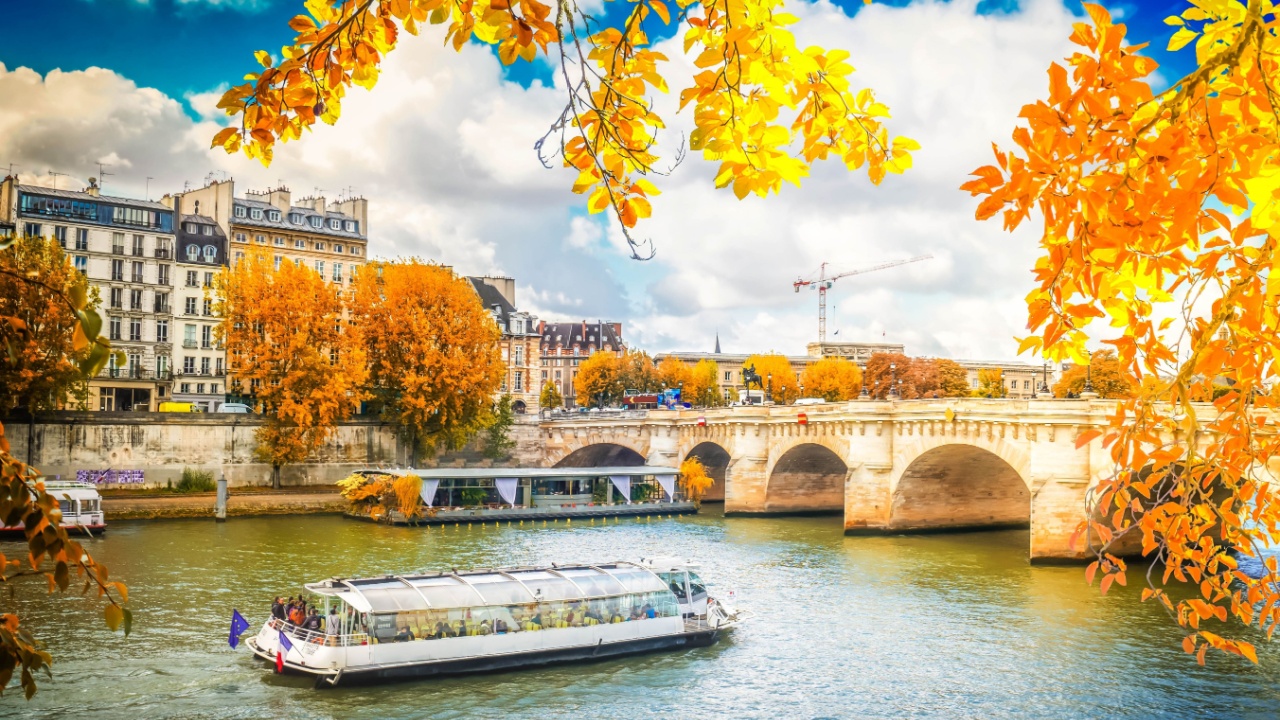 A river cruise boat travels under an arch bridge surrounded by autumn trees and historic buildings on a sunny day.