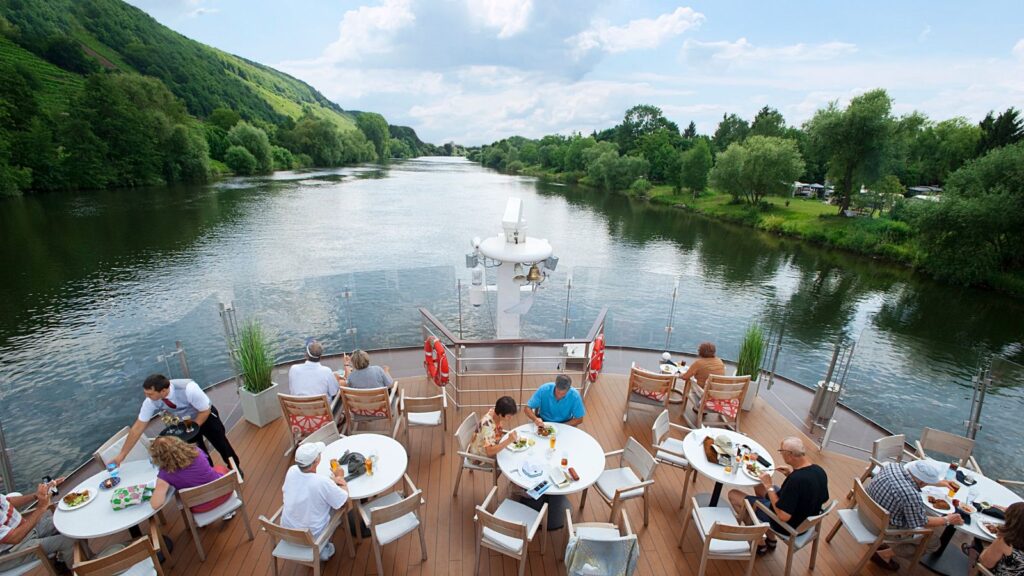 People dining on a boat's deck, cruising along a river surrounded by lush green trees and hilly scenery.