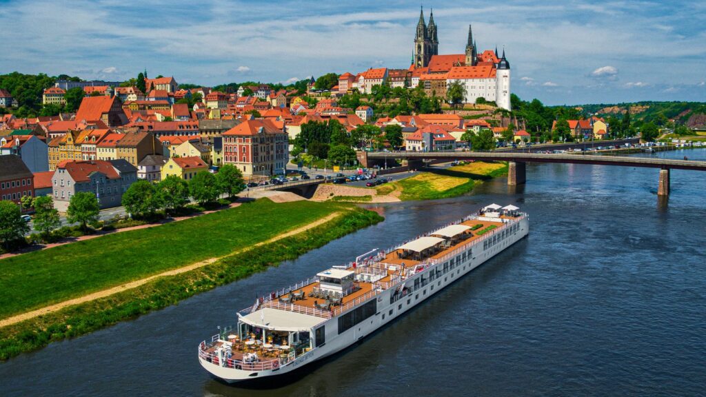 A white river cruise boat sails on a river with a historic town and cathedral on a hill in the background.