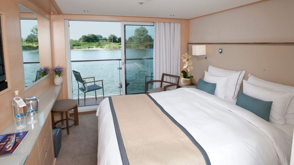 A cozy hotel room with a large bed, glass doors overlooking a balcony and a serene river view in the background.