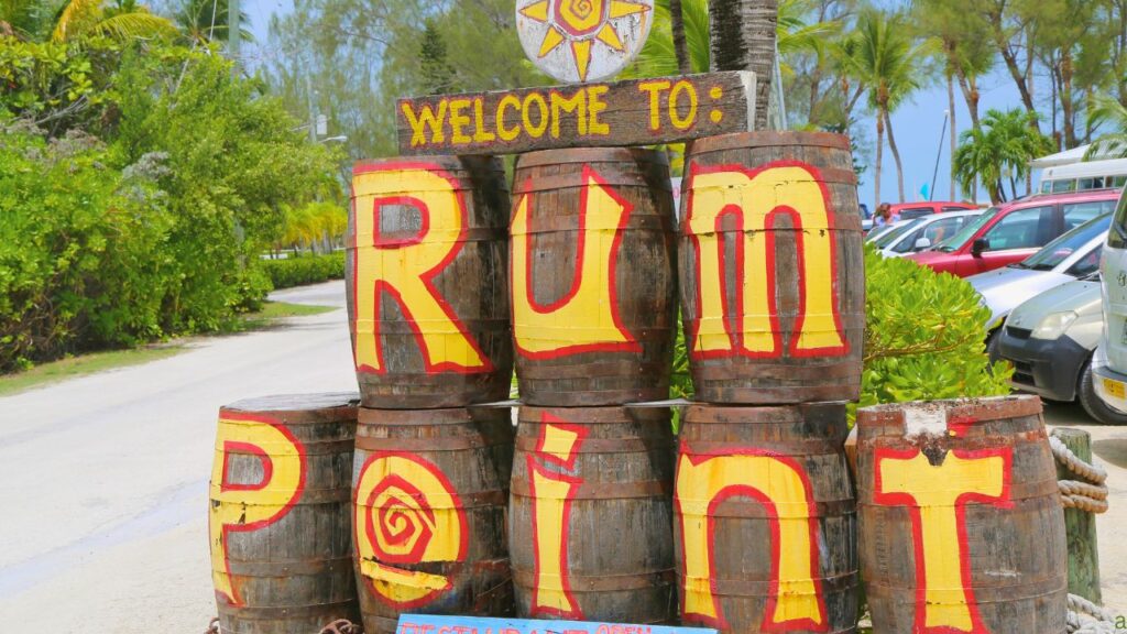 Sign made from barrels reading "Welcome to Rum Point" with tropical background and cars parked nearby.