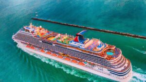 Aerial view of a large, colorful cruise ship sailing in blue-green waters near a long, narrow breakwater.