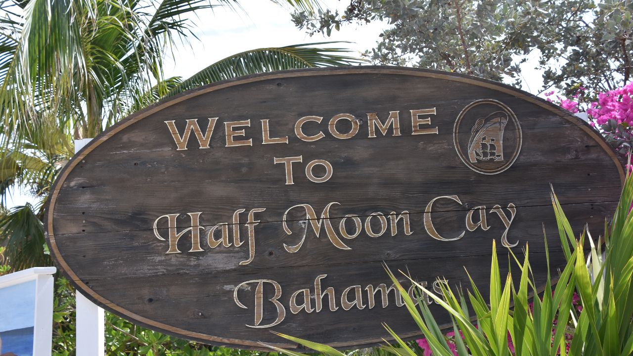 Wooden sign reading "Welcome to Half Moon Cay Bahamas" surrounded by palm trees and tropical plants.