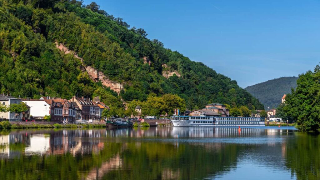 A scenic riverside view with a cruise ship docked by a quaint town and lush green hills.