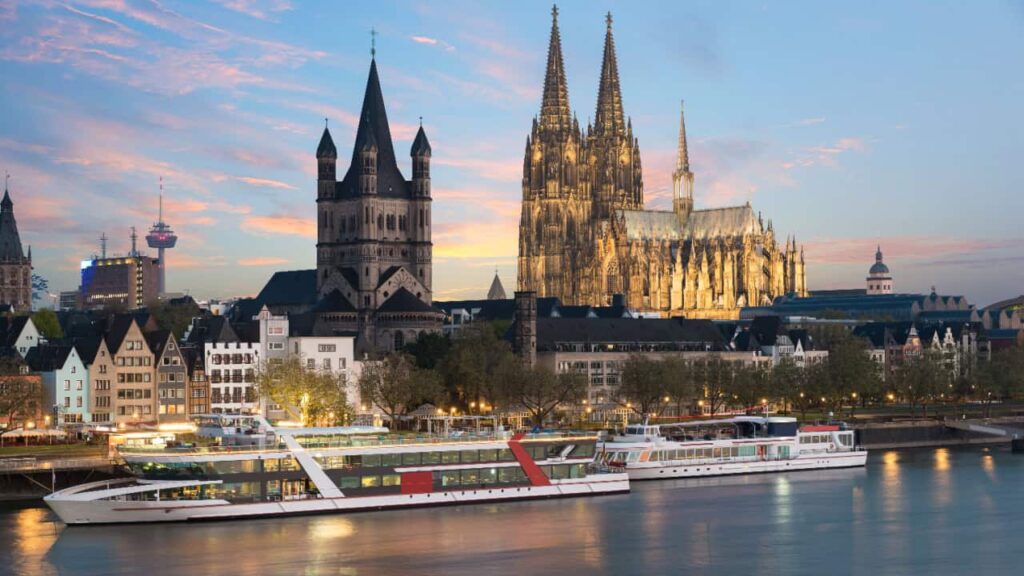 A riverside view of Cologne with the illuminated Cologne Cathedral and boats on the water at sunset.