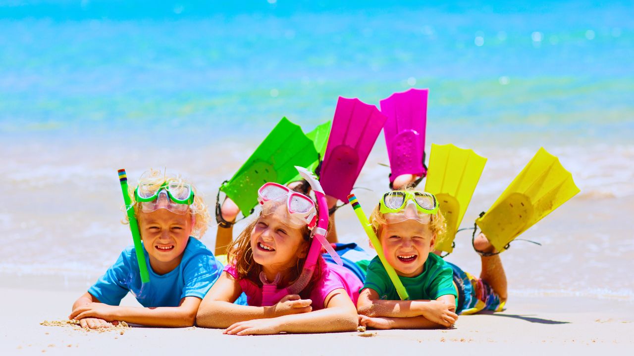 Three children lying on the beach, wearing colorful snorkeling gear and flippers, with the ocean in the background.