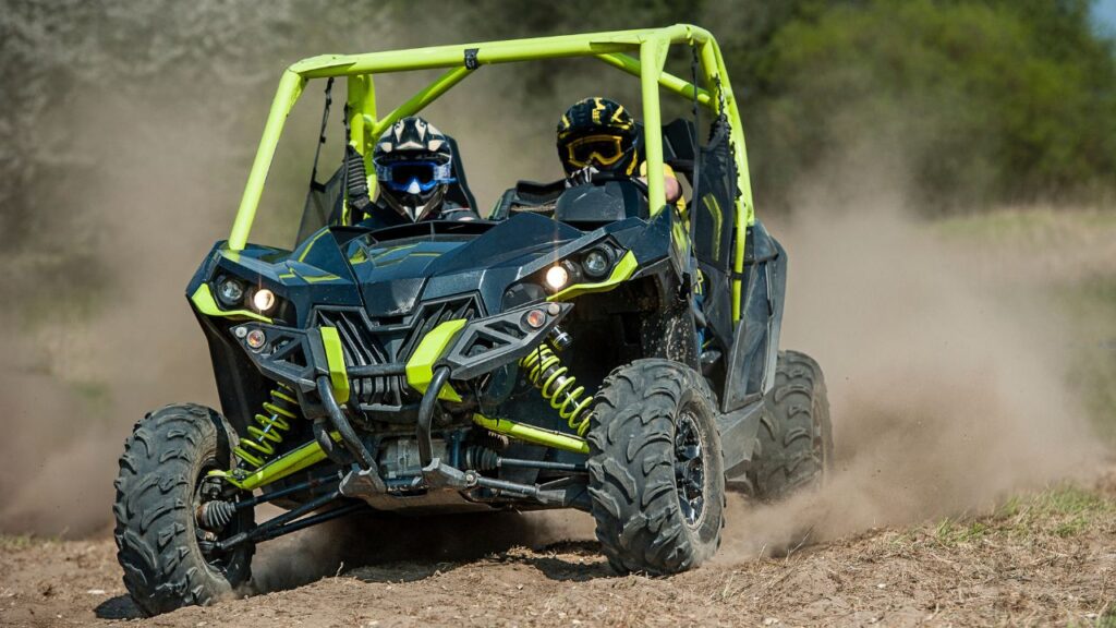 Two people in helmets ride a green and black off-road vehicle on a dusty trail.