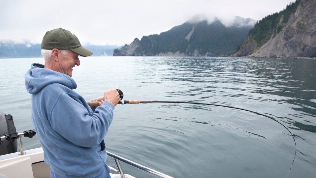 Man fishing on a boat in a calm, misty sea with a mountainous background and cloudy sky.