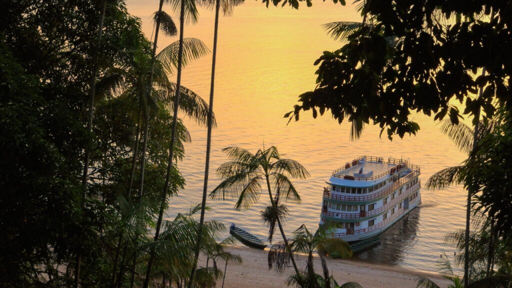 A boat is docked by a sandy beach surrounded by palm trees at sunset.