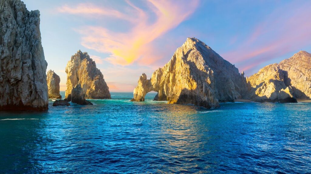 Rocky cliffs at the edge of a calm sea under a sky with pink and orange clouds at sunset.