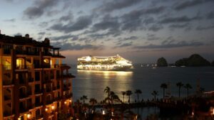 A illuminated cruise ship sails at dusk near a beachside resort with palm trees and several boats on the water.