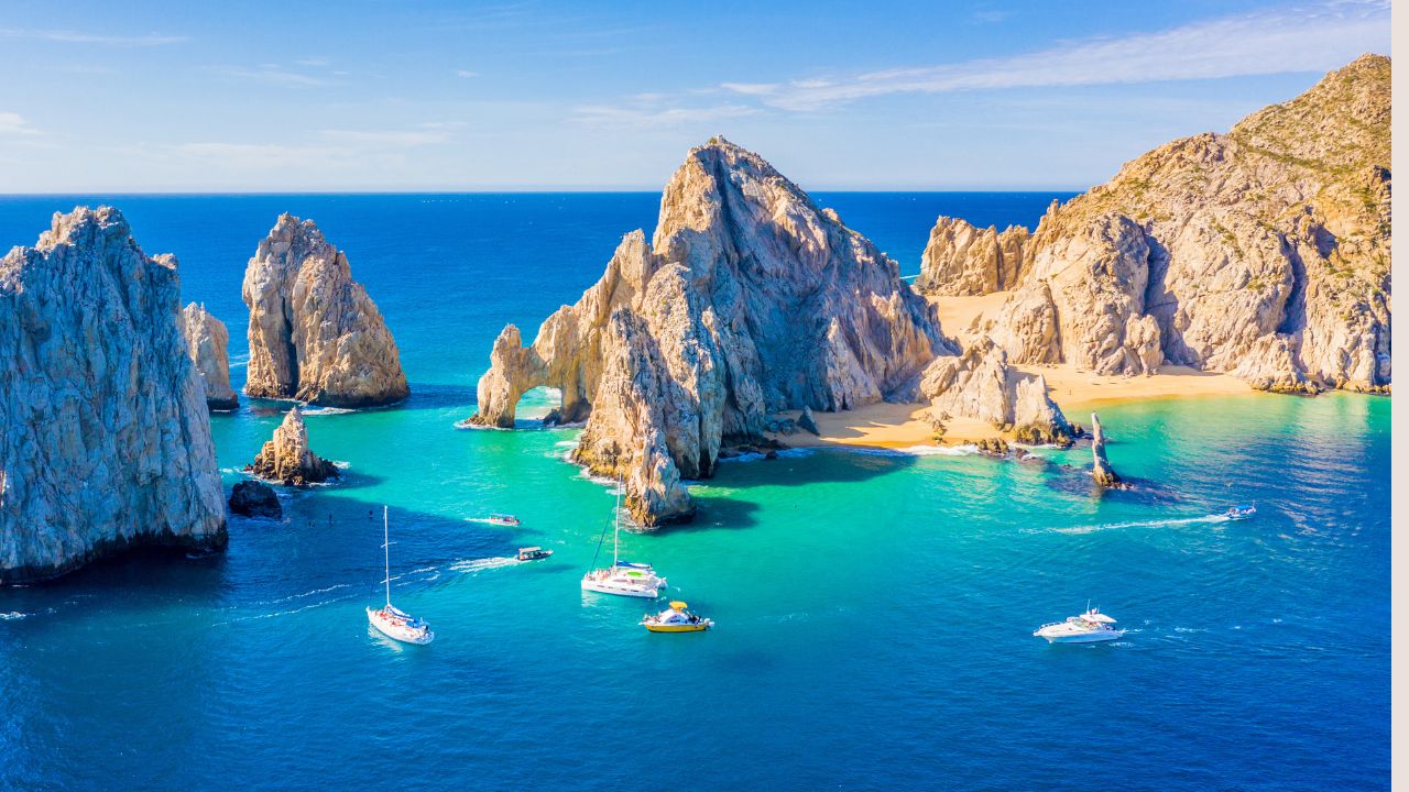 Aerial view of boats near rocky arch formations and turquoise waters at a sunny beach.