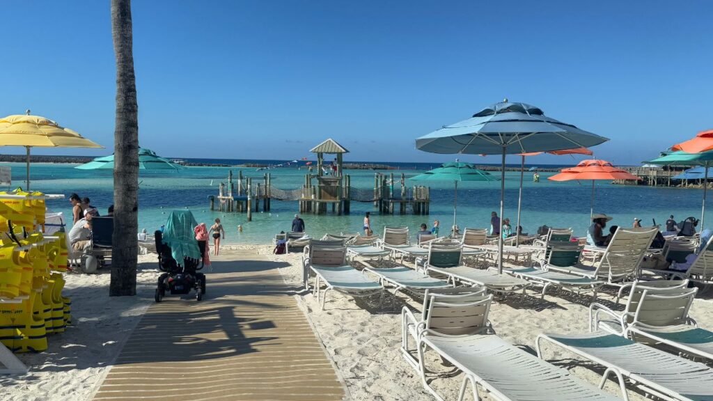 A sunny beach scene with lounge chairs, umbrellas, and people near a clear, calm ocean under a bright blue sky.