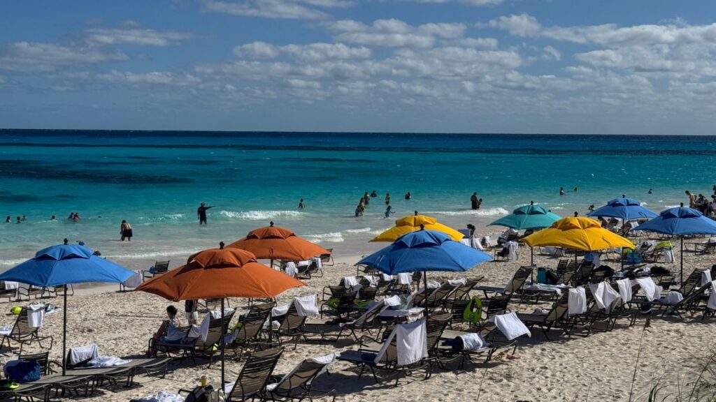 A beach scene with colorful umbrellas, lounge chairs, and people swimming in the ocean under a partly cloudy sky.