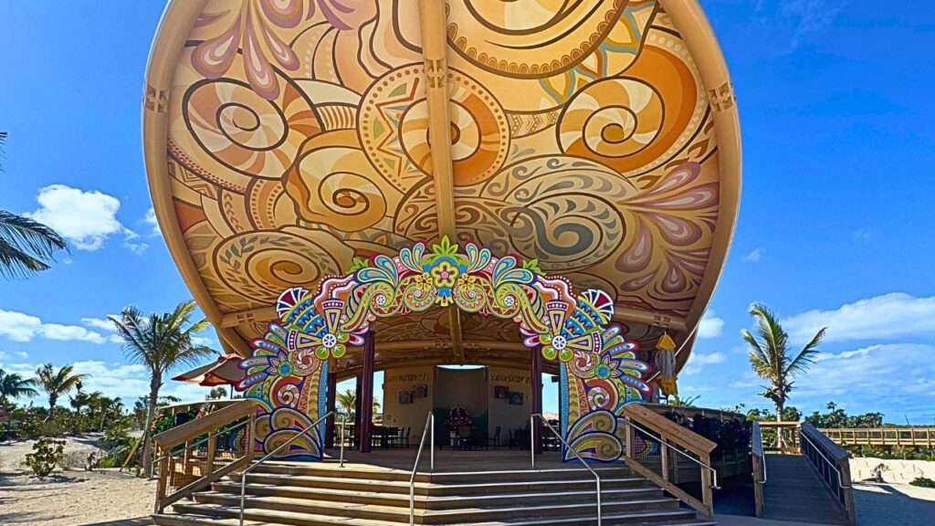 Colorful, intricately patterned dome structure with wooden steps, set against a blue sky and palm trees.