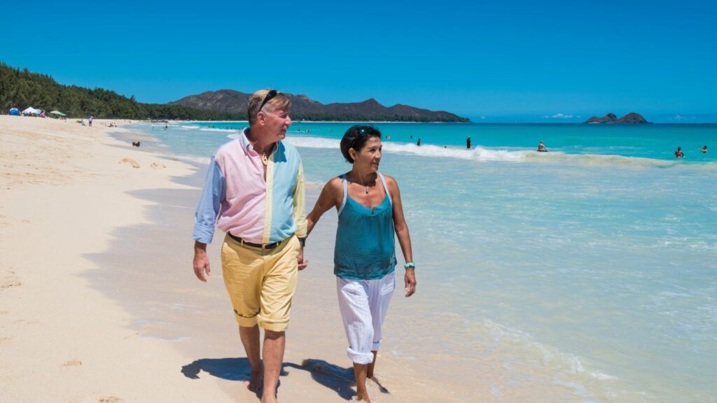 An older couple walks hand in hand along a sunny beach, with clear blue skies and gentle waves in the background.