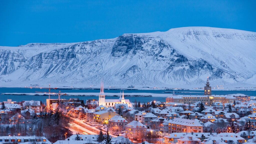 Snowy Reykjavik cityscape at dusk, with illuminated buildings and a mountain backdrop.