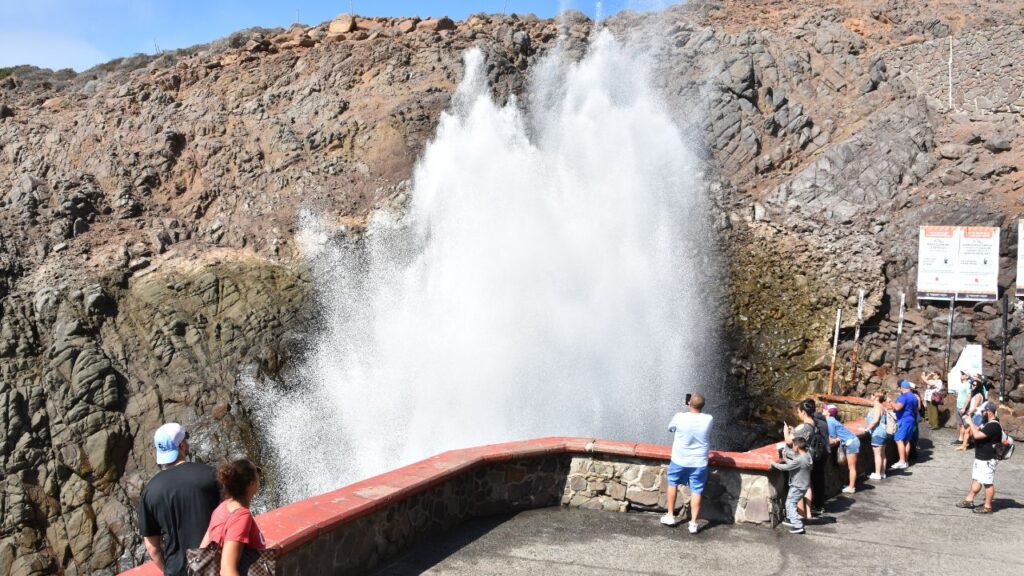 People watching a large ocean geyser spray water against a rocky cliff.