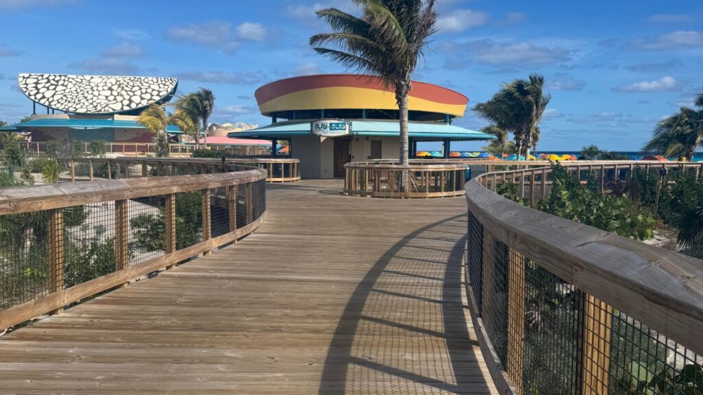 A wooden boardwalk leads to a colorful circular building by the beach, with palm trees and a blue sky above.